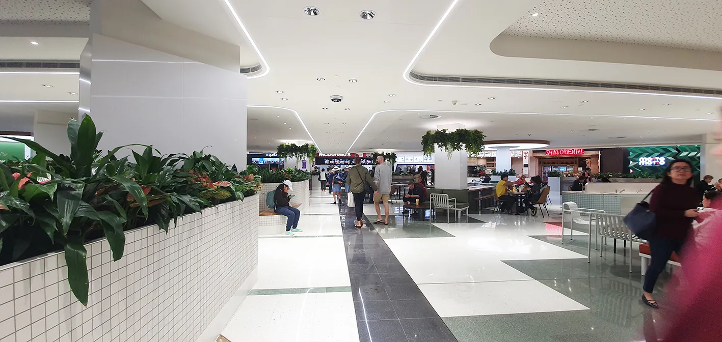 Indoor mall food court with people dining and walking, surrounded by eateries and plant decor.