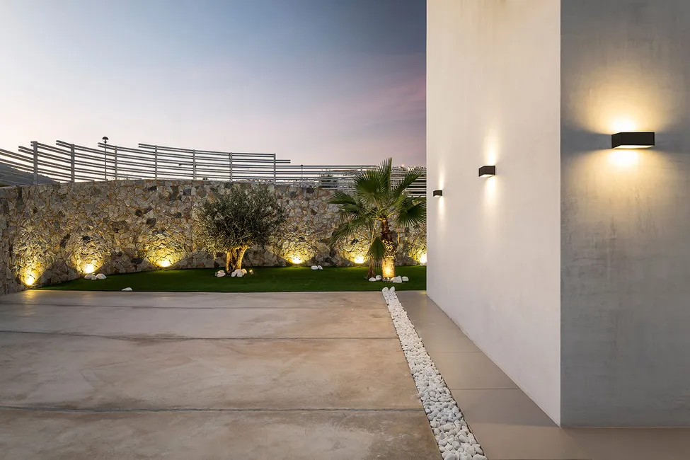 Evening view of a modern outdoor space with lighting on a stone wall and sleek wall sconces.