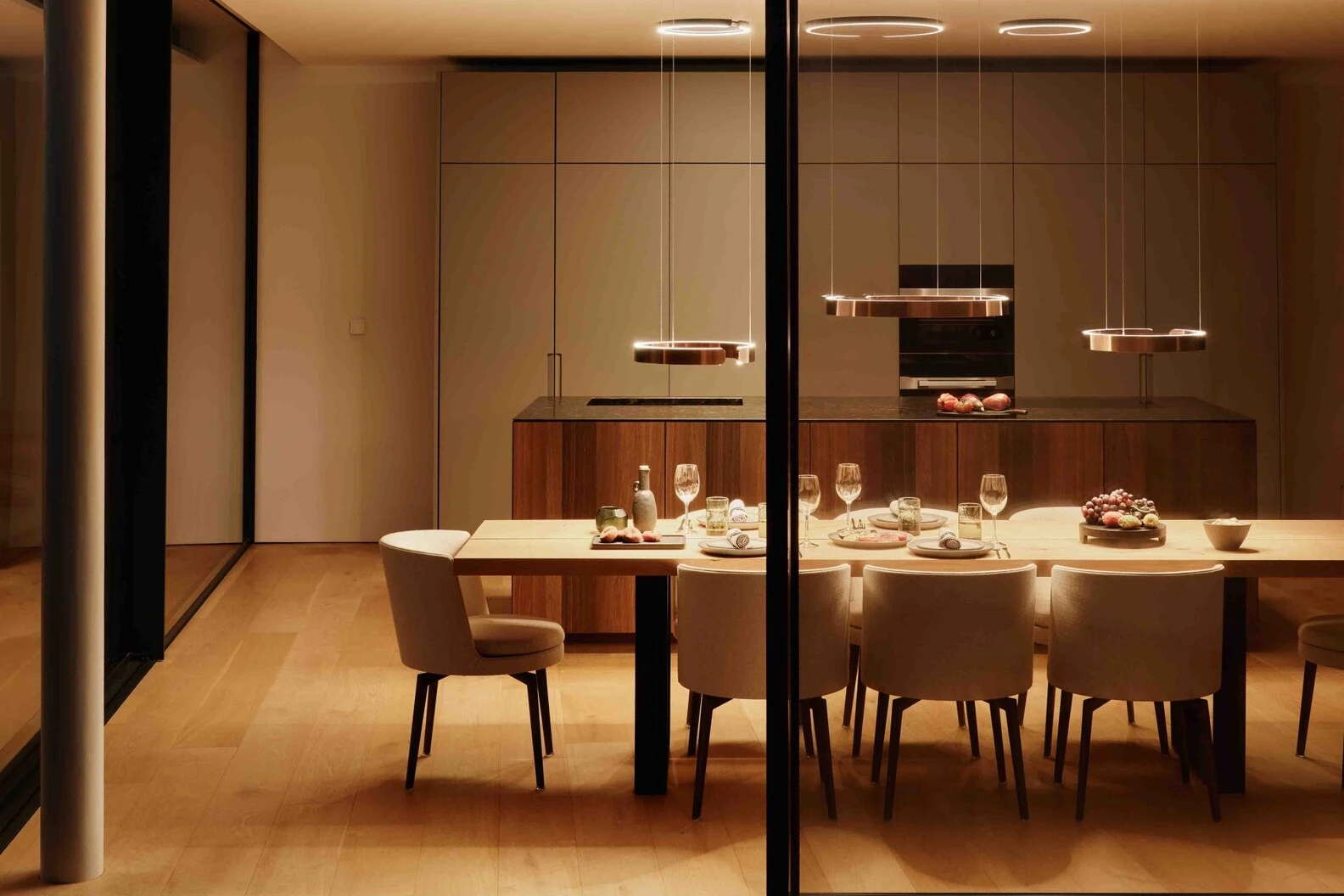 Modern dining room with wooden table, chairs, and pendant lights, viewed through glass.