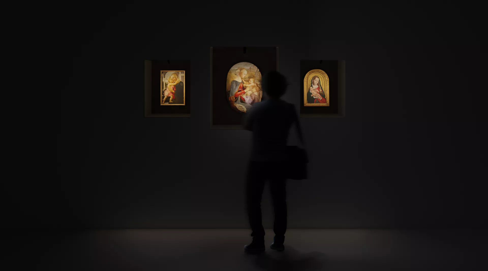 A person gazes thoughtfully at various paintings displayed in a dimly lit room, immersed in the art around them.