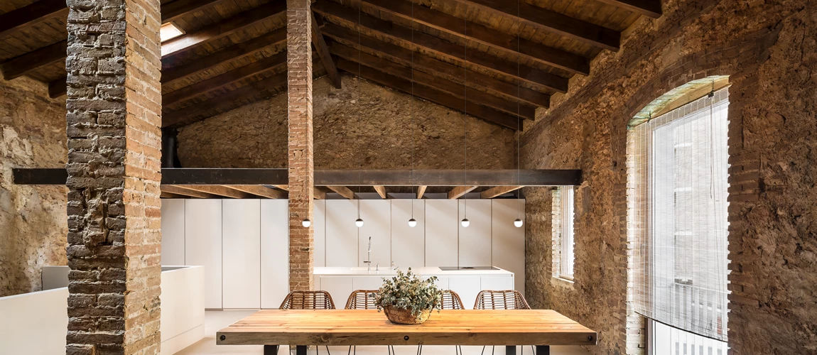 Modern kitchen with exposed brick walls and wooden beams, featuring a large table and minimalist decor.